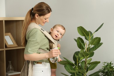 Mother spraying houseplant with water while holding her child in sling (baby carrier) at home