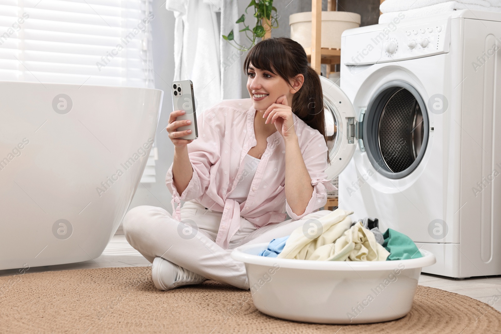 Photo of Happy young housewife with laundry using smartphone near washing machine at home