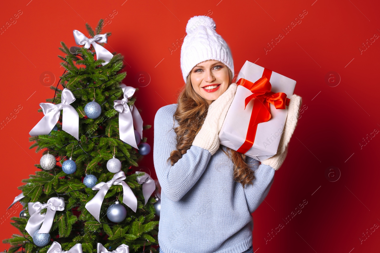 Photo of Beautiful young woman in hat and mittens with gift box near Christmas tree on color background