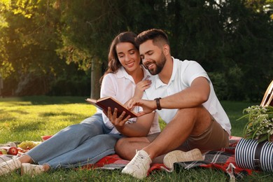 Photo of Happy young couple reading book on plaid in park. Summer picnic