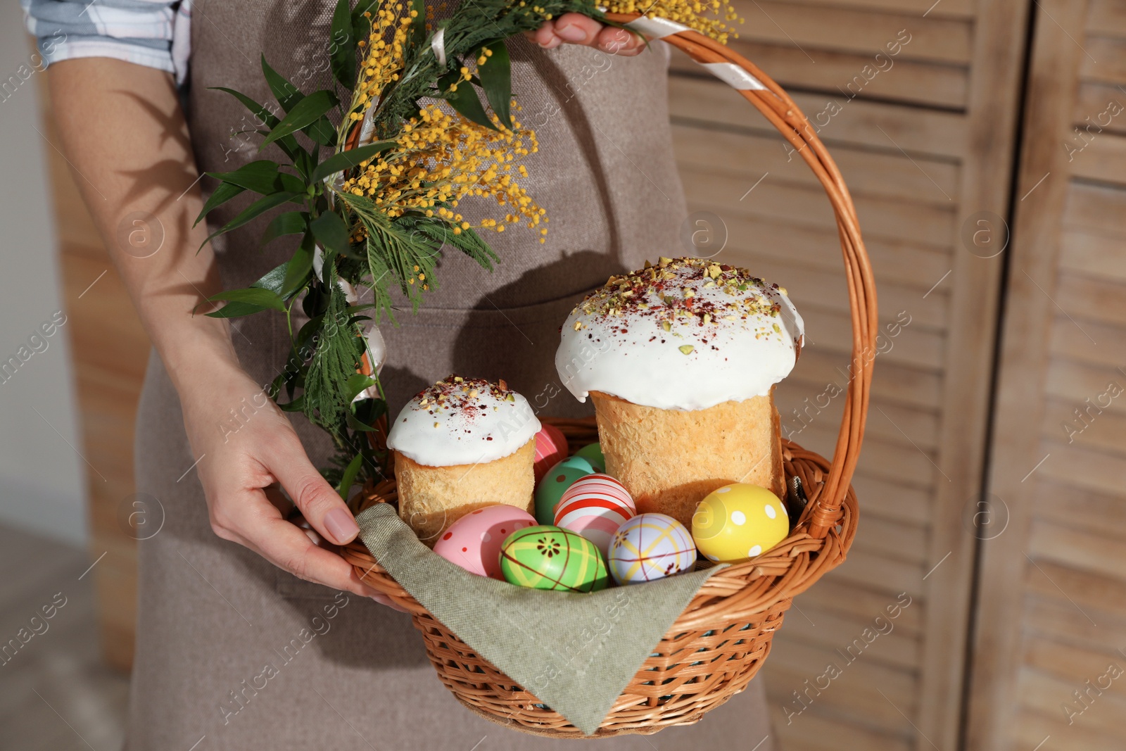 Photo of Woman holding basket with traditional Easter cakes, dyed eggs and flowers indoors, closeup