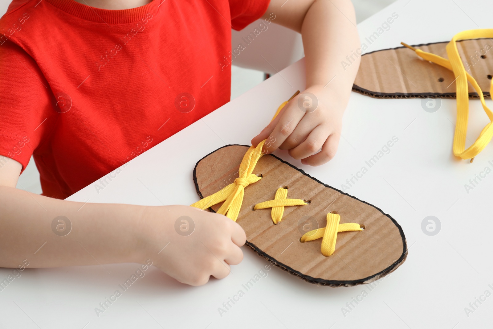 Photo of Little boy tying shoe lace using training cardboard template at white table, closeup