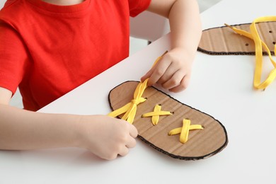 Little boy tying shoe lace using training cardboard template at white table, closeup