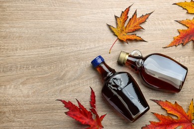 Photo of Glass bottles of tasty maple syrup and dry leaves on wooden table, flat lay. Space for text