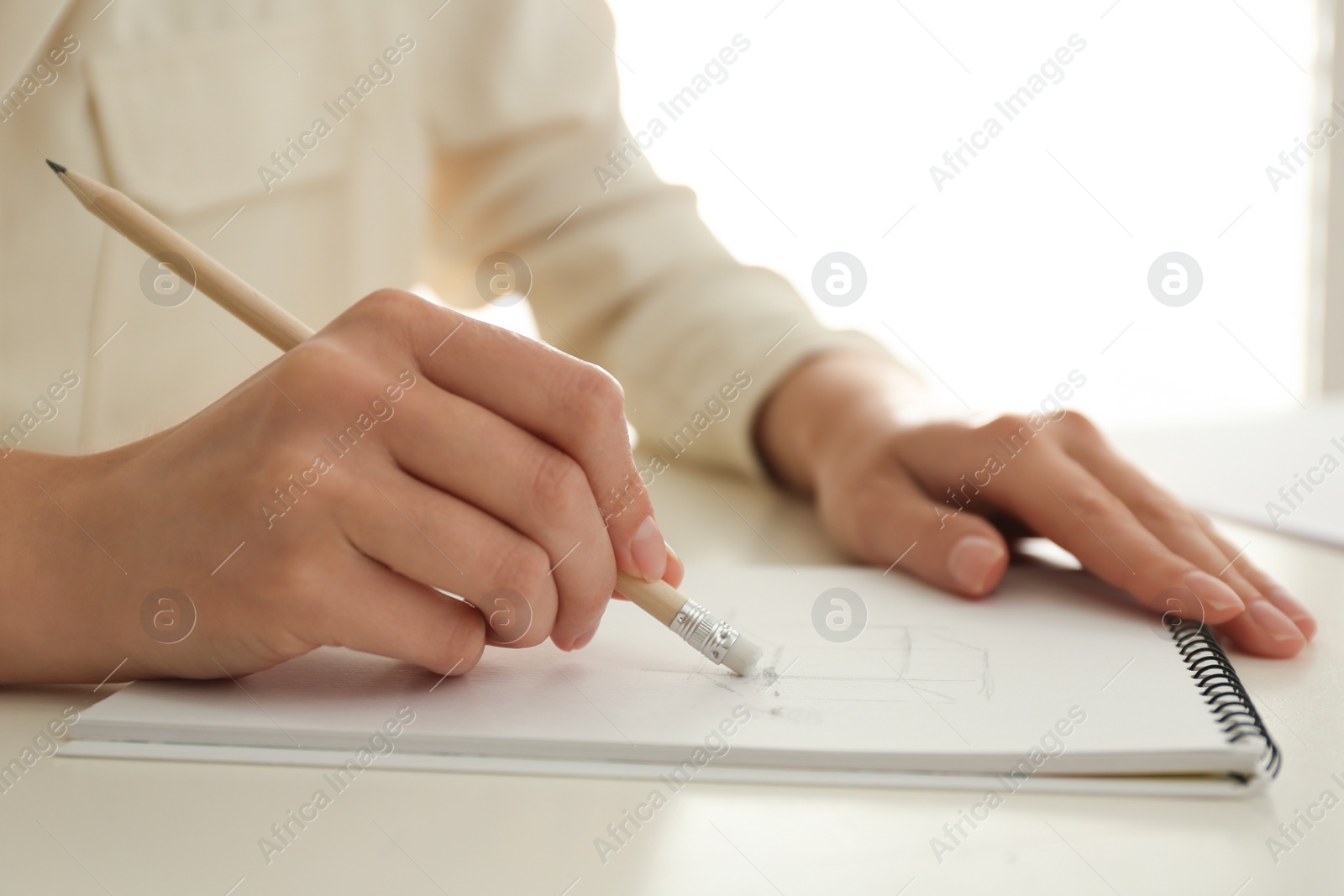 Photo of Woman correcting picture in notepad with pencil eraser at white table, closeup