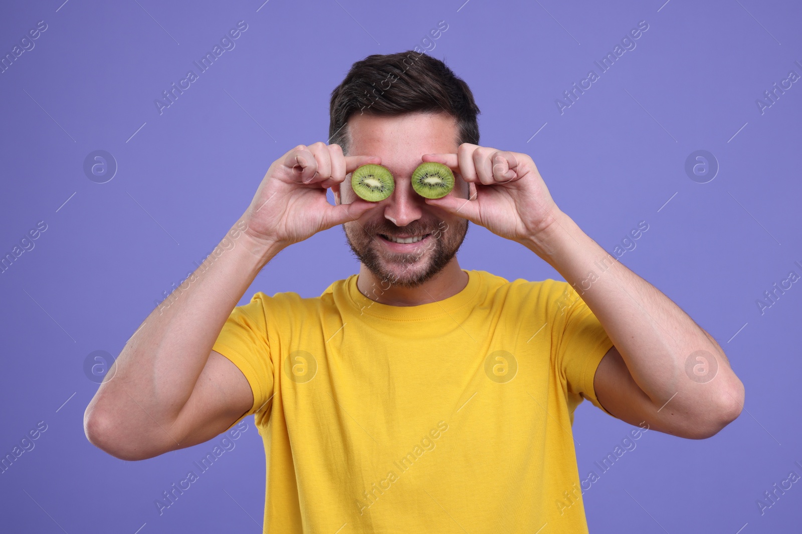 Photo of Man covering his eyes with halves of kiwi on purple background