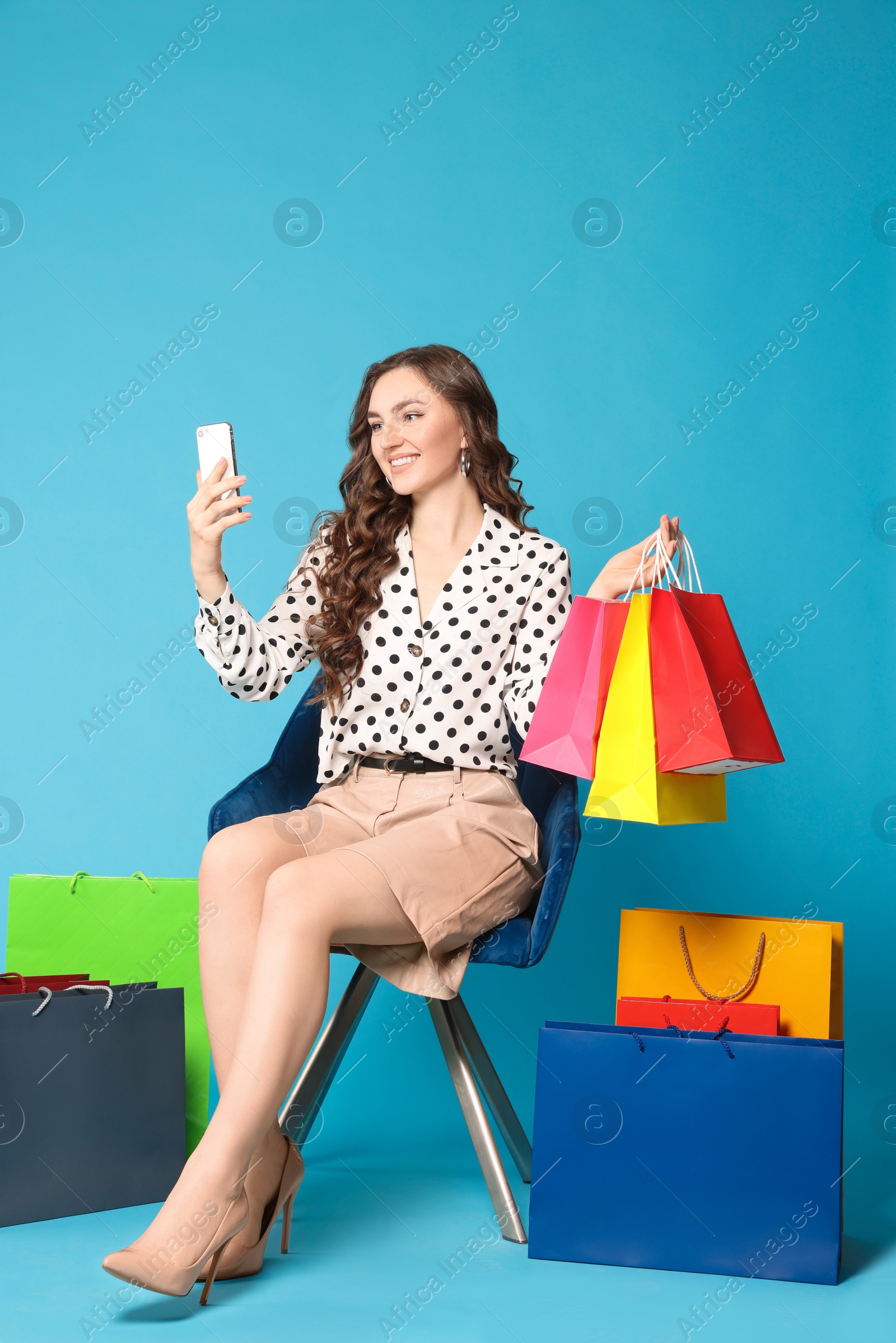 Photo of Happy woman taking selfie while holding colorful shopping bags on armchair against light blue background