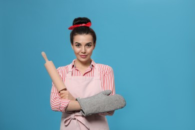 Photo of Young housewife in oven glove holding roller pin on light blue background, space for text