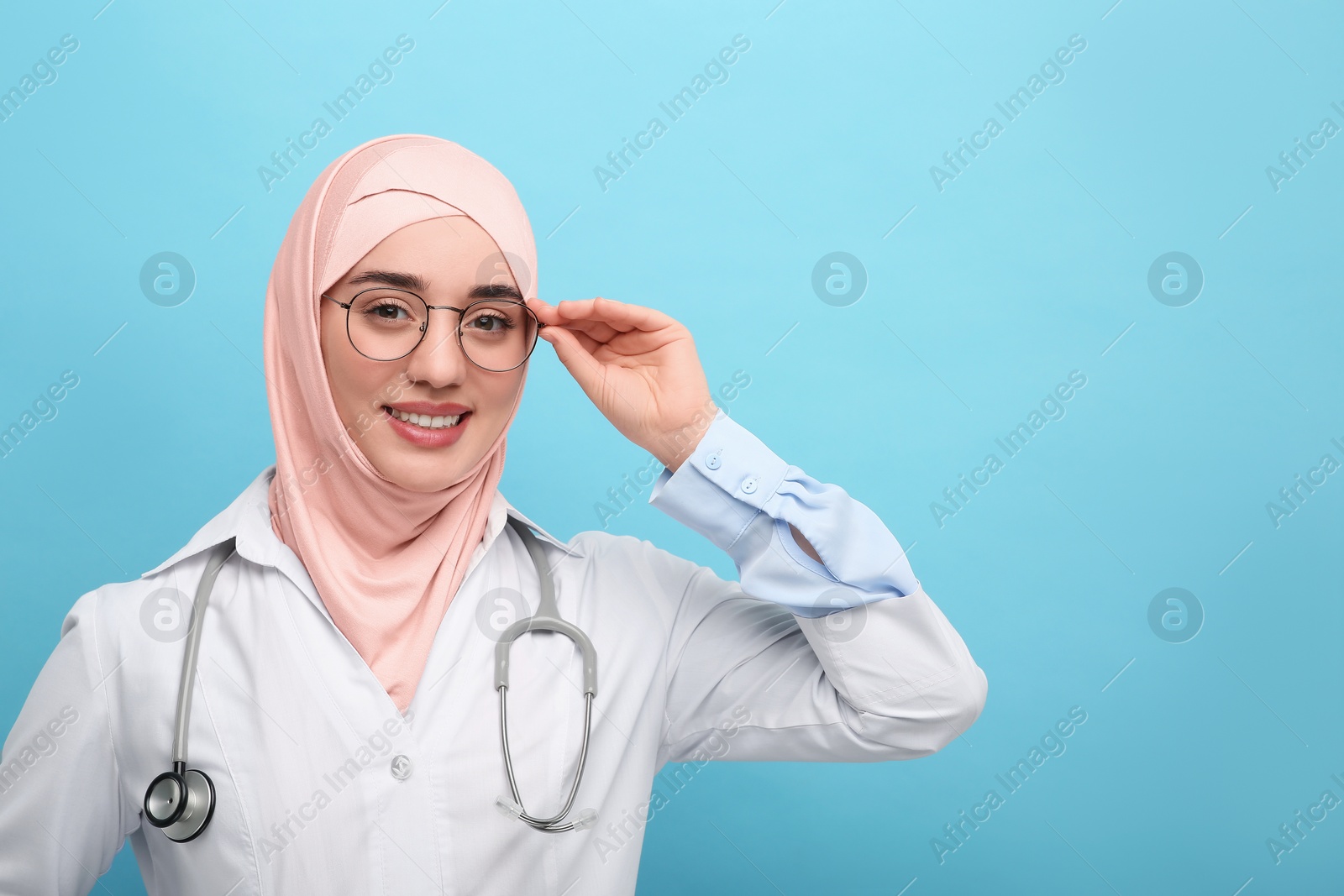 Photo of Muslim woman wearing hijab and medical uniform with stethoscope on light blue background, space for text