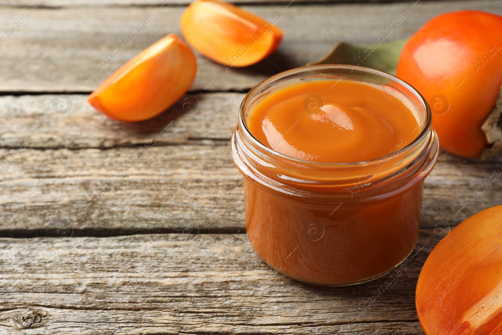 Photo of Delicious persimmon jam in glass jar and fresh fruits on wooden table, closeup. Space for text