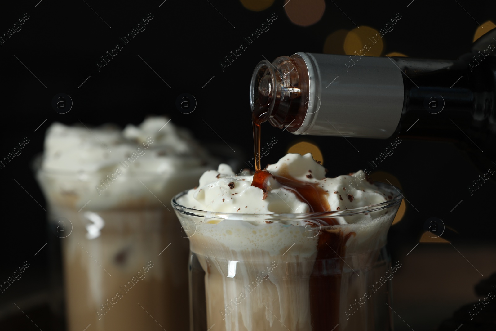 Photo of Pouring syrup into glass of tasty iced coffee against blurred lights, closeup