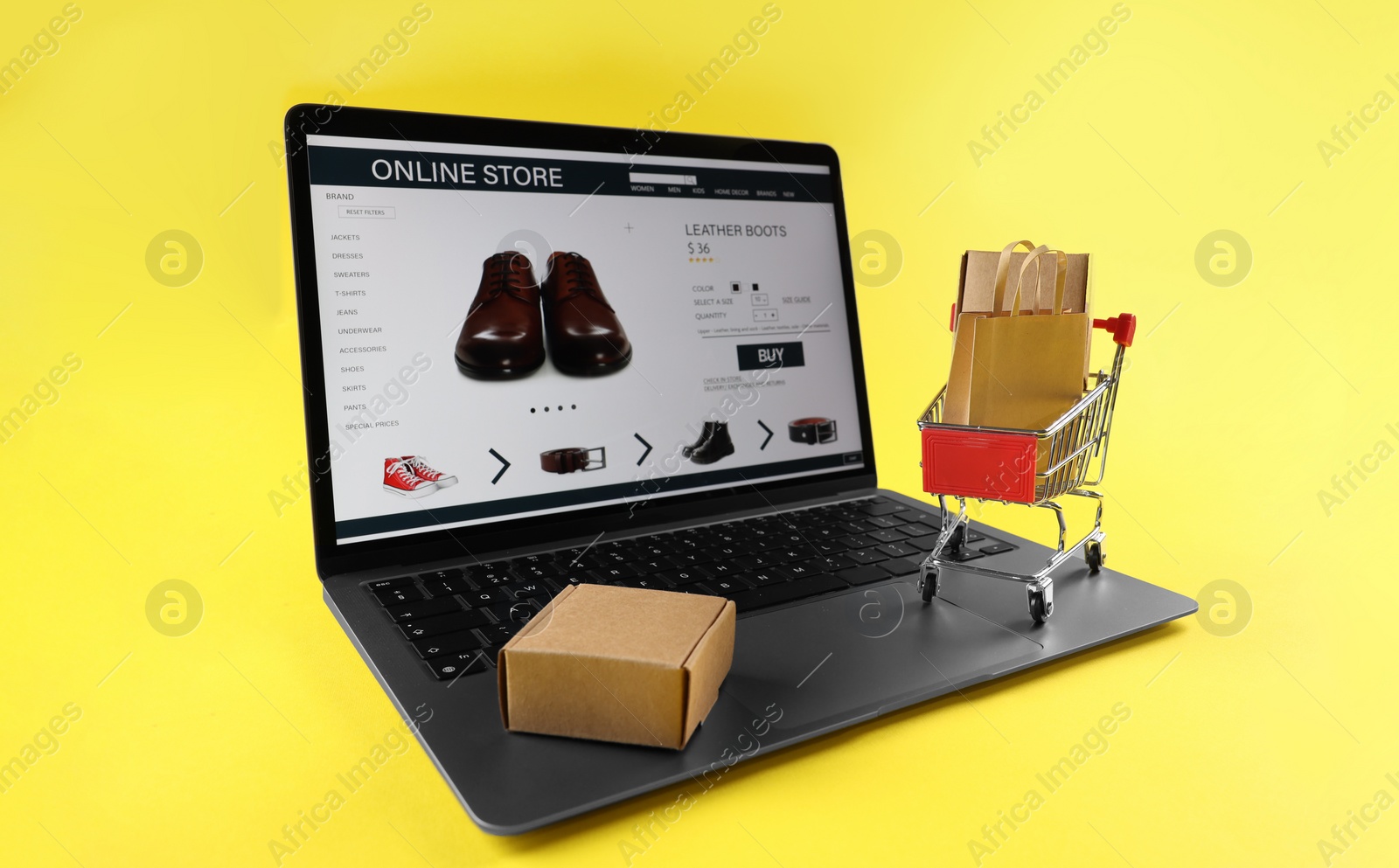 Photo of Online store. Laptop, mini shopping cart and purchases on yellow background