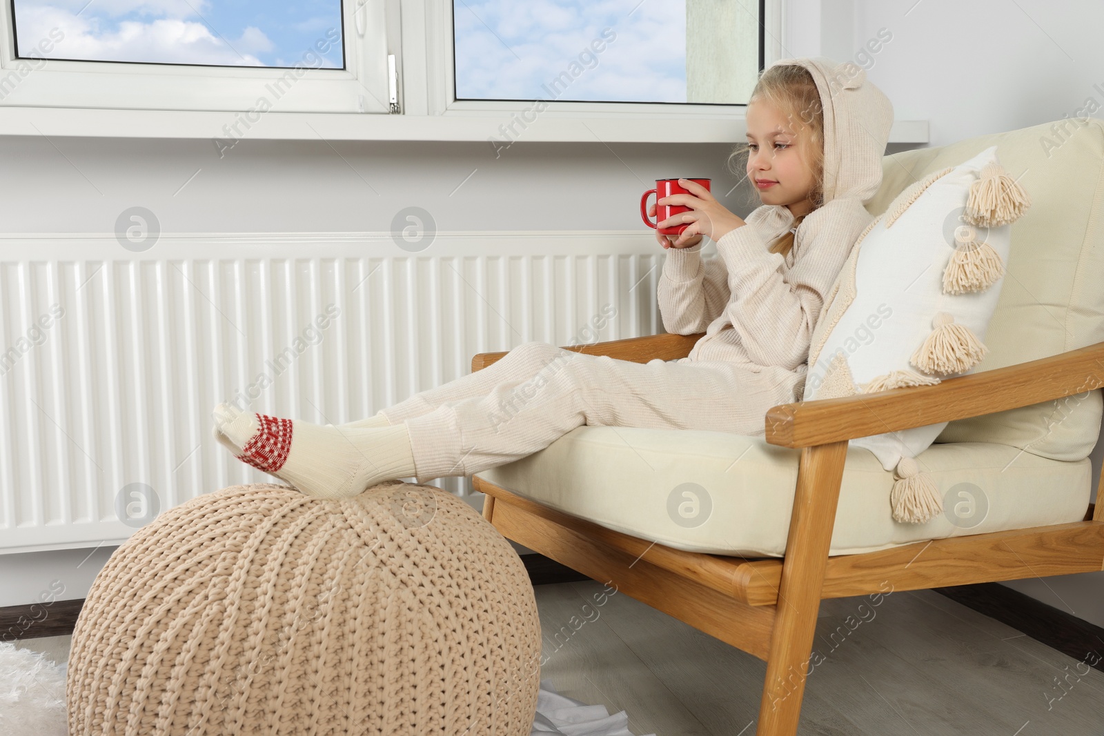 Photo of Little girl with cup of hot drink near heating radiator indoors