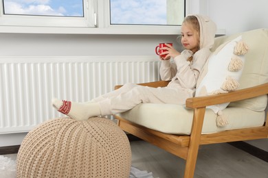 Little girl with cup of hot drink near heating radiator indoors