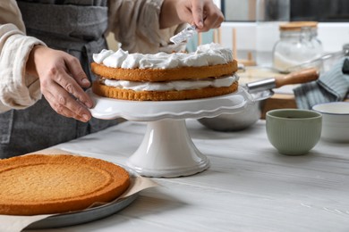 Photo of Woman smearing sponge cake with cream at white wooden table in kitchen, closeup
