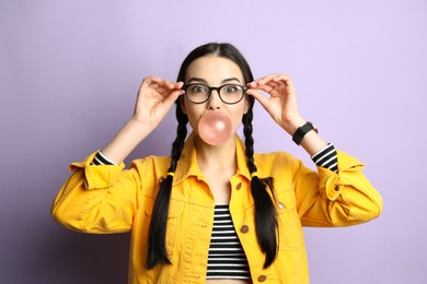 Photo of Fashionable young woman with braids blowing bubblegum on lilac background