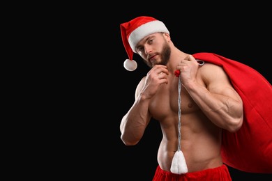 Muscular young man in Santa hat holding bag with presents on black background, space for text