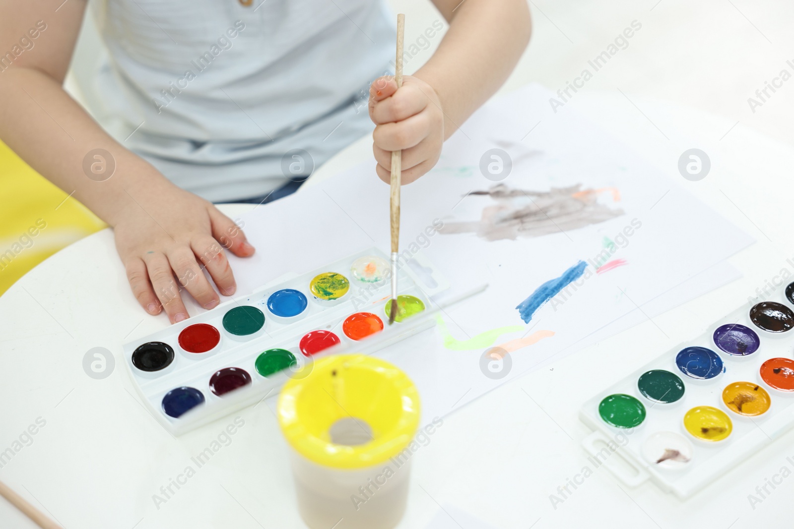 Photo of Little boy painting with brush and watercolor at white table, closeup