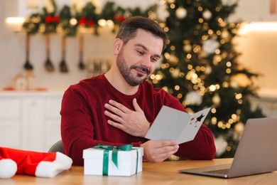 Celebrating Christmas online with exchanged by mail presents. Man reading greeting card during video call on laptop at home