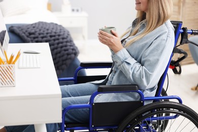 Photo of Woman in wheelchair with cup of drink using computer at home, closeup