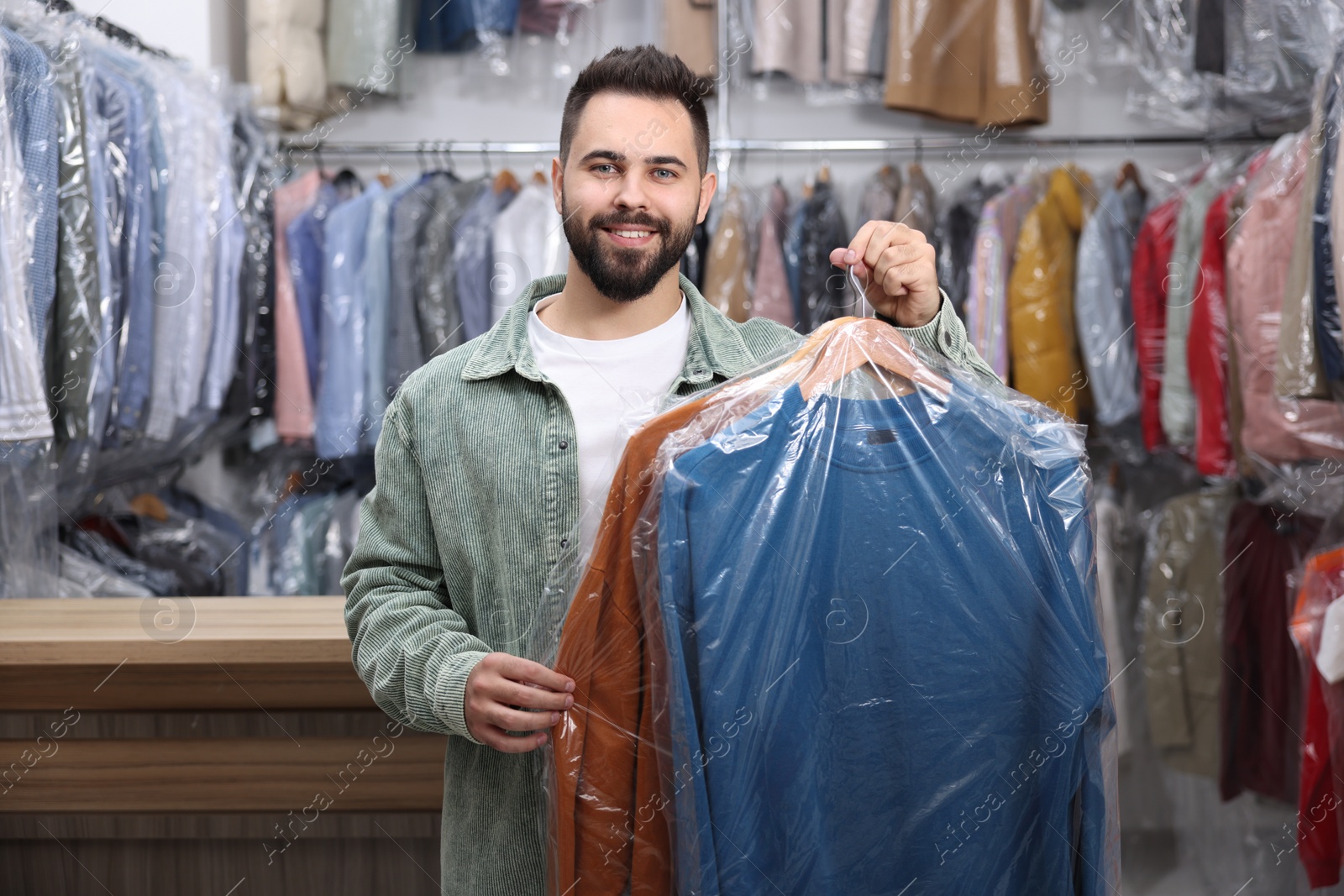 Photo of Dry-cleaning service. Happy man holding hangers with clothes in plastic bags indoors
