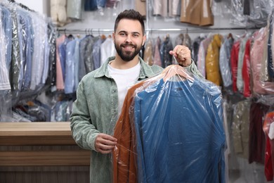 Photo of Dry-cleaning service. Happy man holding hangers with clothes in plastic bags indoors