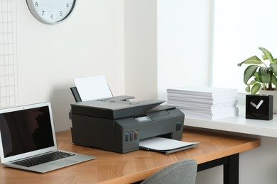 Modern printer with paper and laptop on wooden table in office