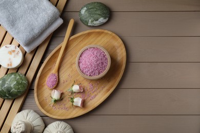 Bowl of pink sea salt, roses, spa stones, herbal massage bags and towels on wooden table, flat lay. Space for text