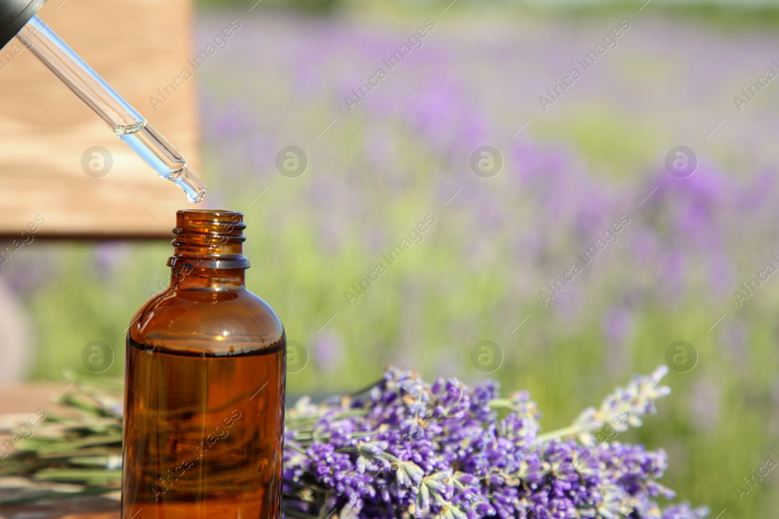 Photo of Dripping essential oil from pipette into bottle near lavender on wooden table outdoors, closeup. Space for text