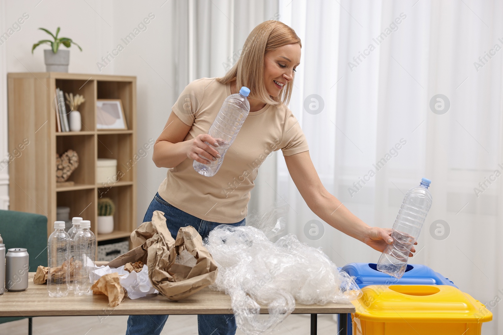 Photo of Garbage sorting. Smiling woman throwing plastic bottle into trash bin in room