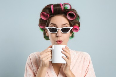 Photo of Beautiful young woman in bathrobe with hair curlers holding cup of drink on light grey background