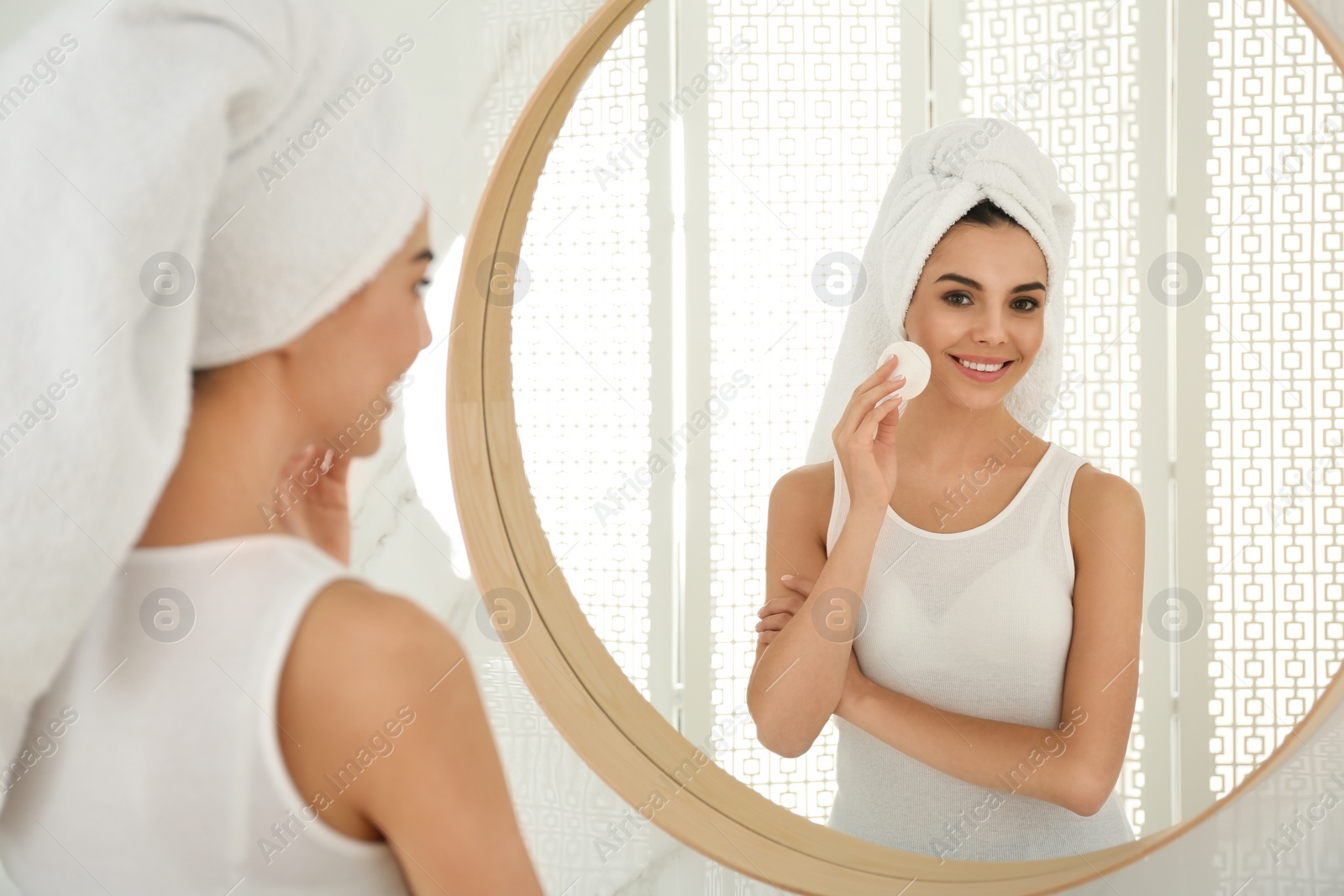 Photo of Happy young woman cleaning face with cotton pad near mirror in bathroom