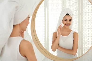 Happy young woman cleaning face with cotton pad near mirror in bathroom