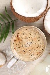 Cup of coffee, halves of coconut, flakes, syrup and green leaves on white wooden table, flat lay