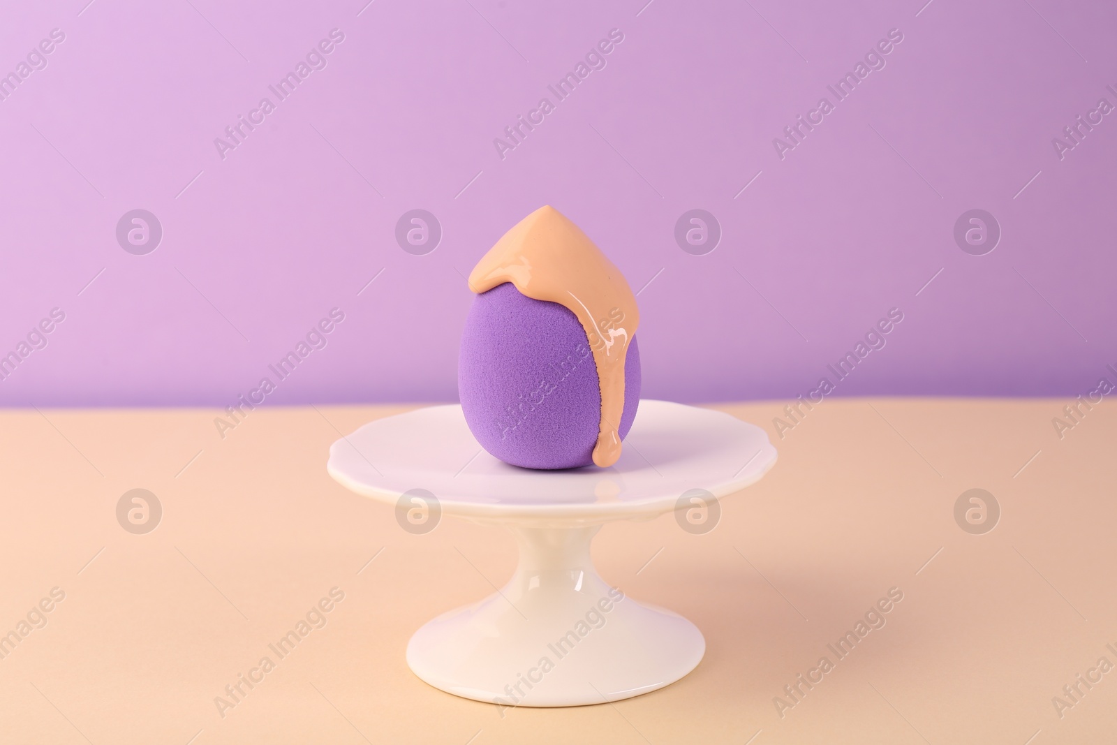 Photo of Stand with makeup sponge and skin foundation on beige table against violet background