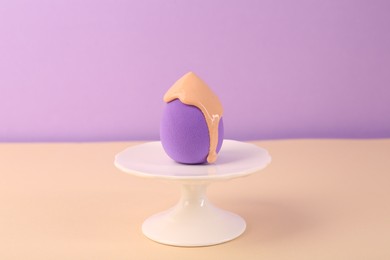 Stand with makeup sponge and skin foundation on beige table against violet background