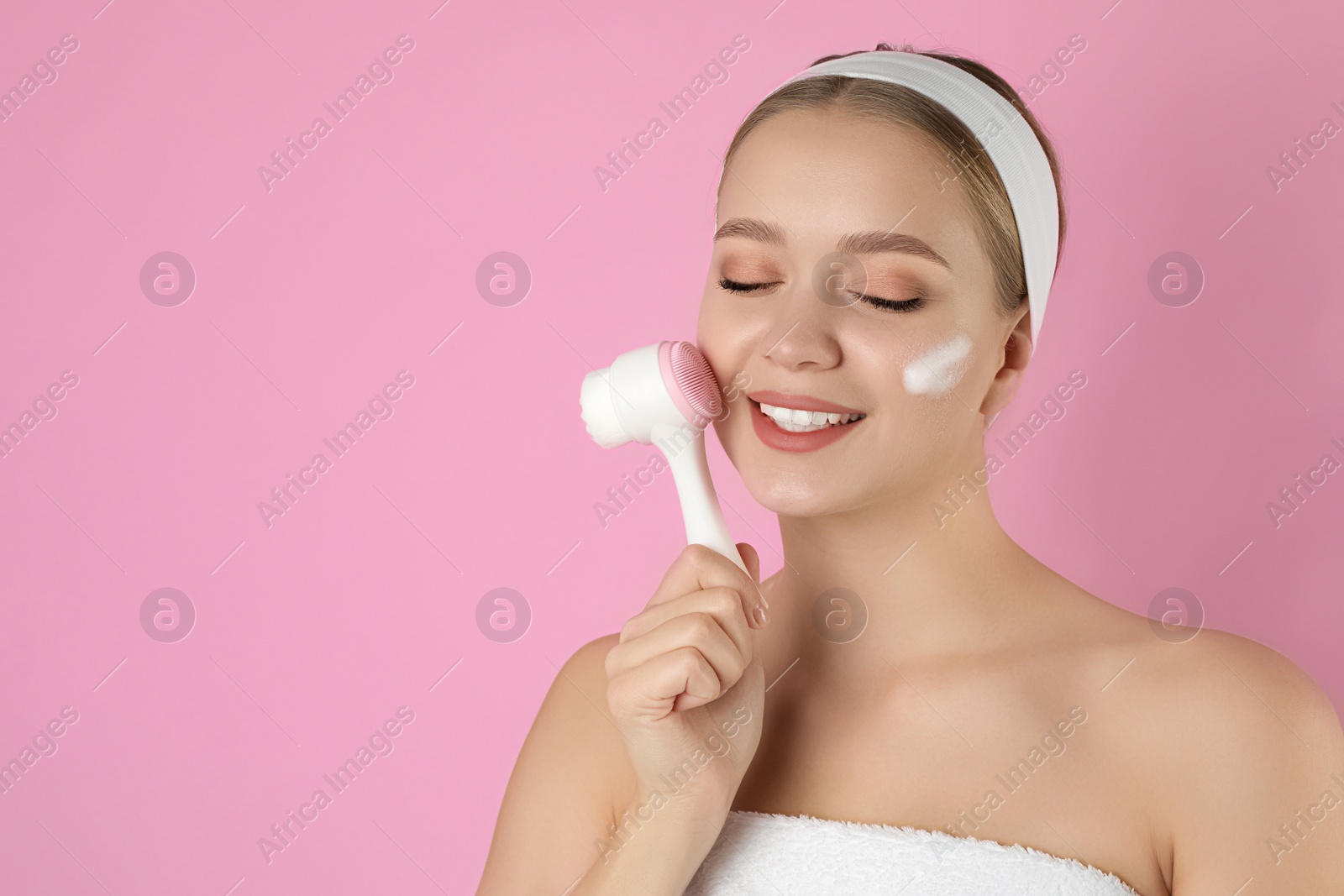 Photo of Young woman washing face with brush and cleansing foam on pink background, space for text. Cosmetic product