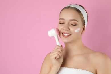 Young woman washing face with brush and cleansing foam on pink background, space for text. Cosmetic product