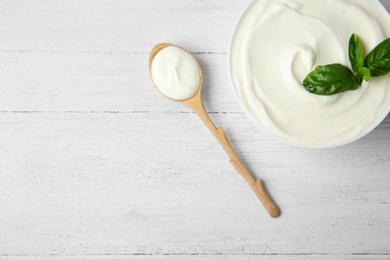 Photo of Bowl of fresh sour cream with basil and spoon on white wooden table, flat lay. Space for text