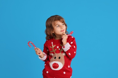 Cute little girl in Christmas sweater holding sweet candy canes against blue background