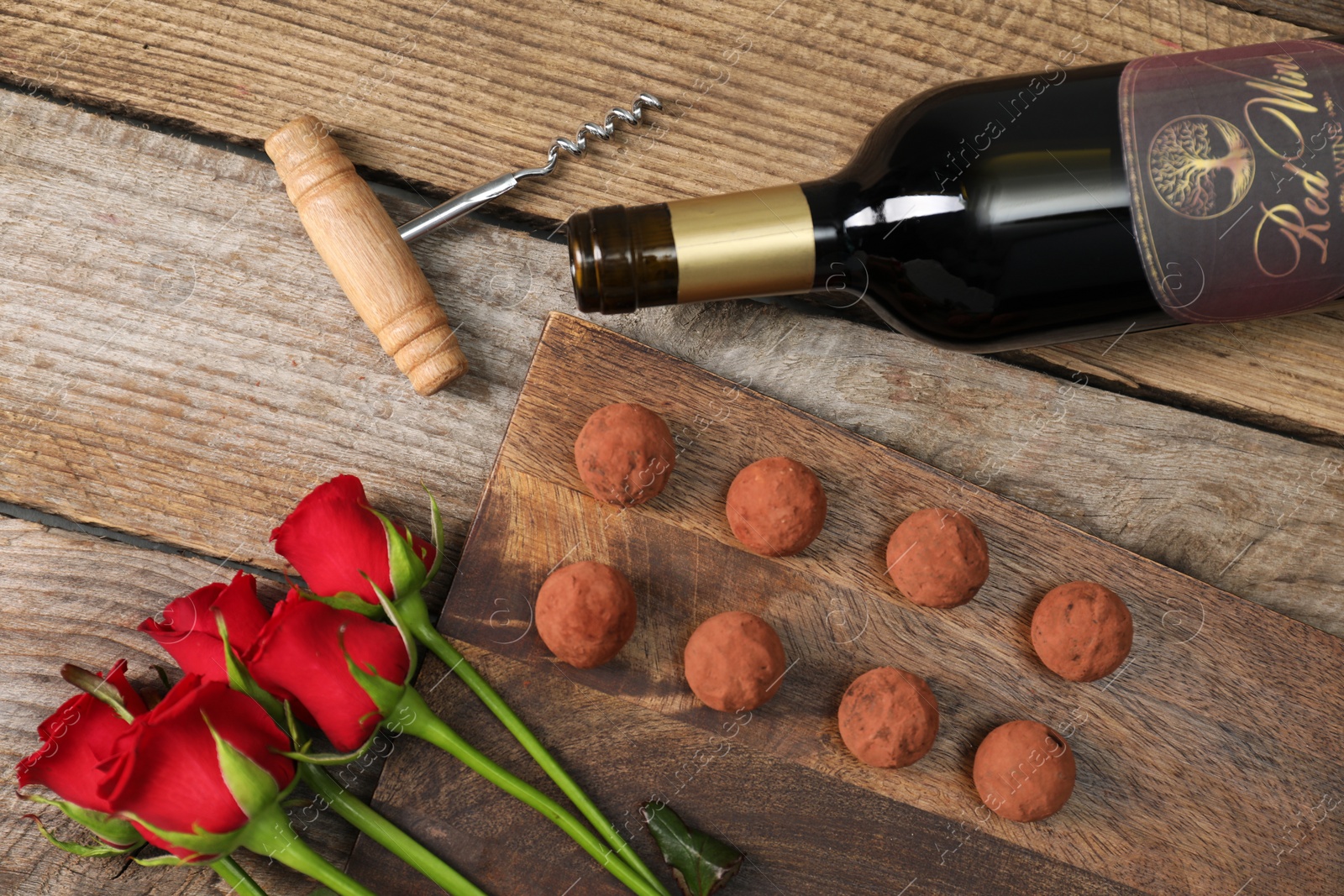 Photo of Bottle of red wine, chocolate truffles, corkscrew and roses on wooden table, flat lay