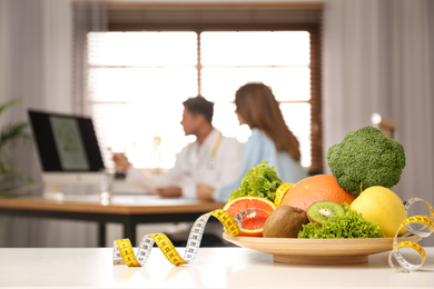 Nutritionist consulting patient at table in clinic, focus on plate with fruits, vegetables and measuring tape