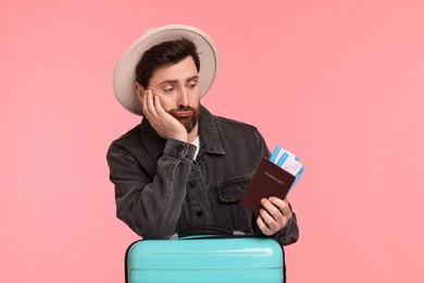 Disappointed man with passport, tickets and suitcase on pink background