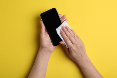 Photo of Woman cleaning mobile phone with antiseptic wipe on yellow background, top view