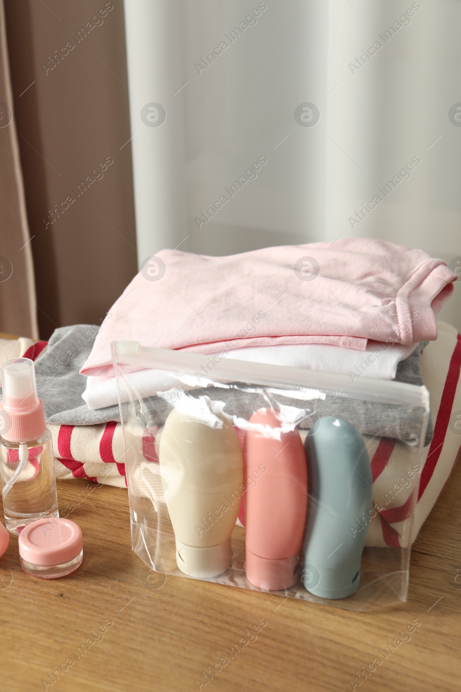 Photo of Cosmetic travel kit. Plastic bag with small containers of personal care products and stack of clothes on wooden table indoors