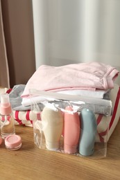 Photo of Cosmetic travel kit. Plastic bag with small containers of personal care products and stack of clothes on wooden table indoors