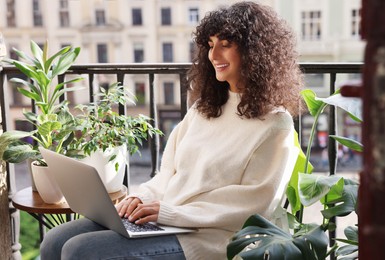 Beautiful young woman using laptop surrounded by green houseplants on balcony