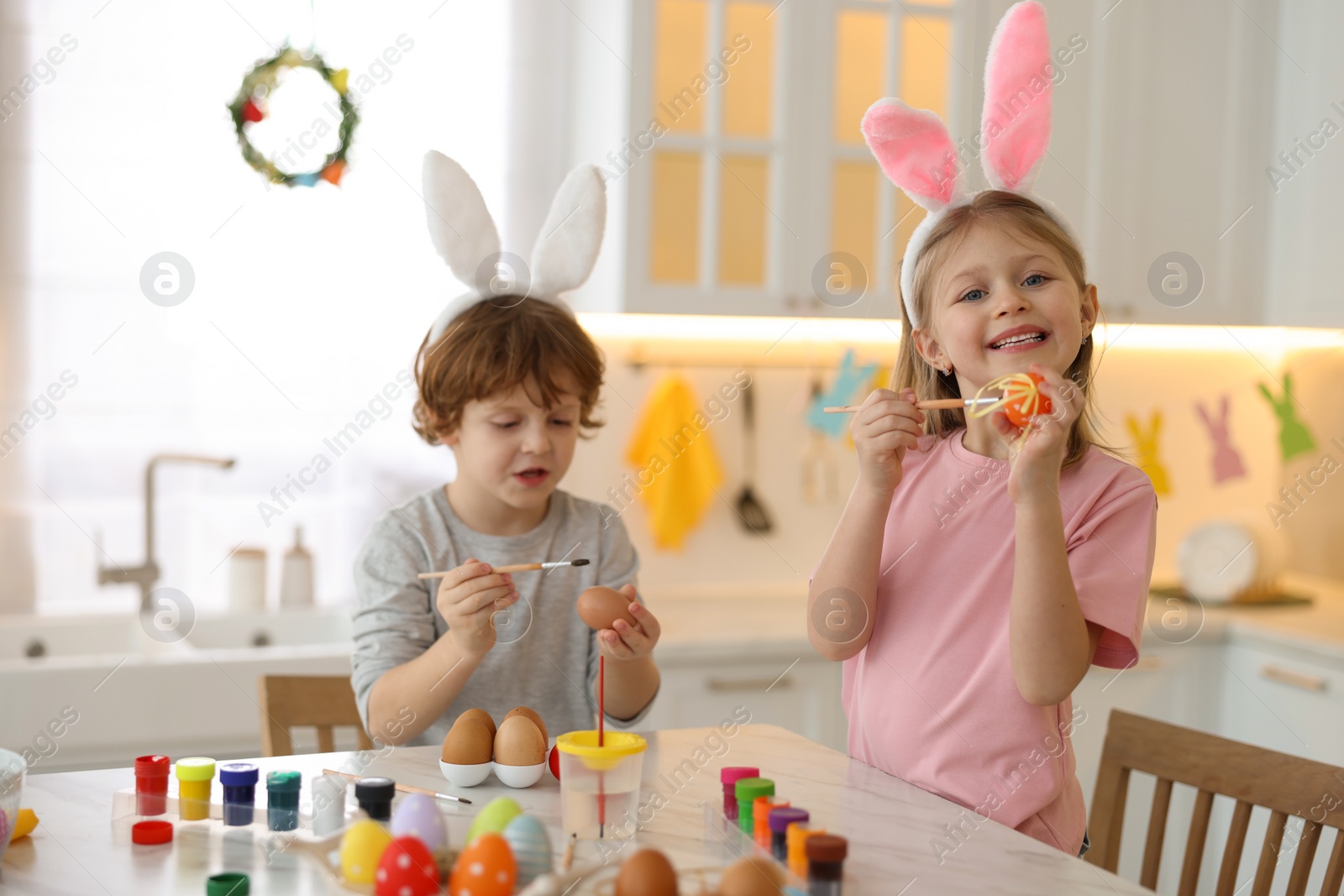 Photo of Easter celebration. Cute children with bunny ears painting eggs at white marble table in kitchen