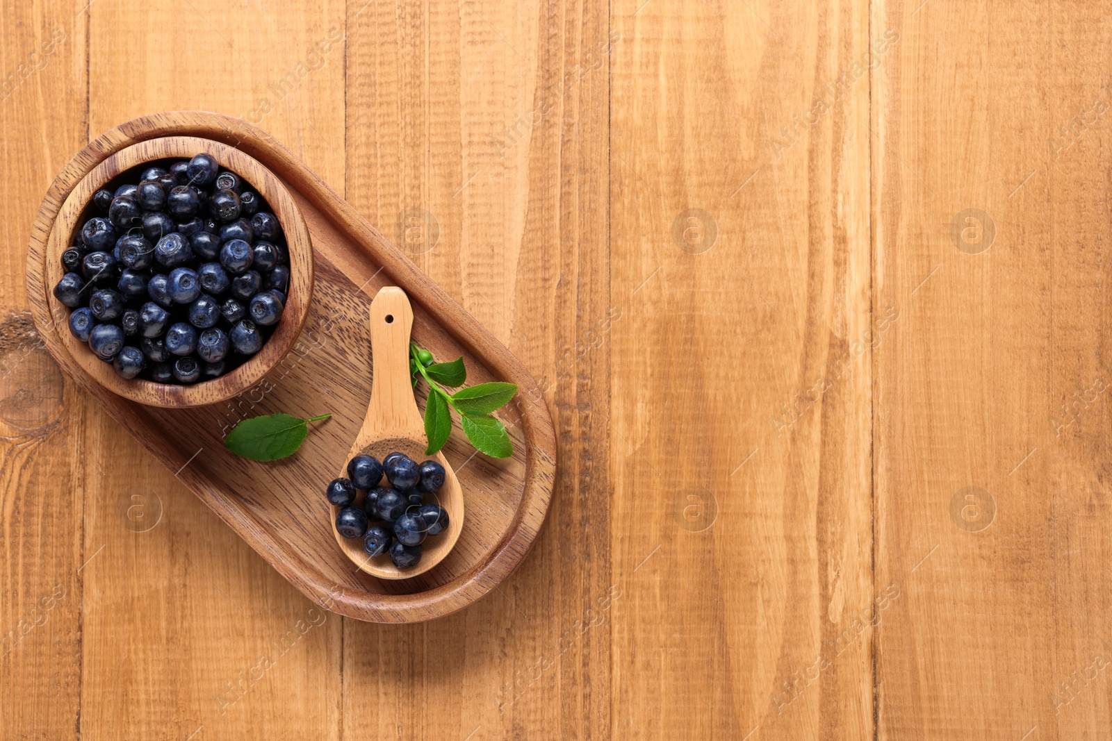 Photo of Bowl, spoon with tasty fresh bilberries and leaves on wooden table, top view. Space for text