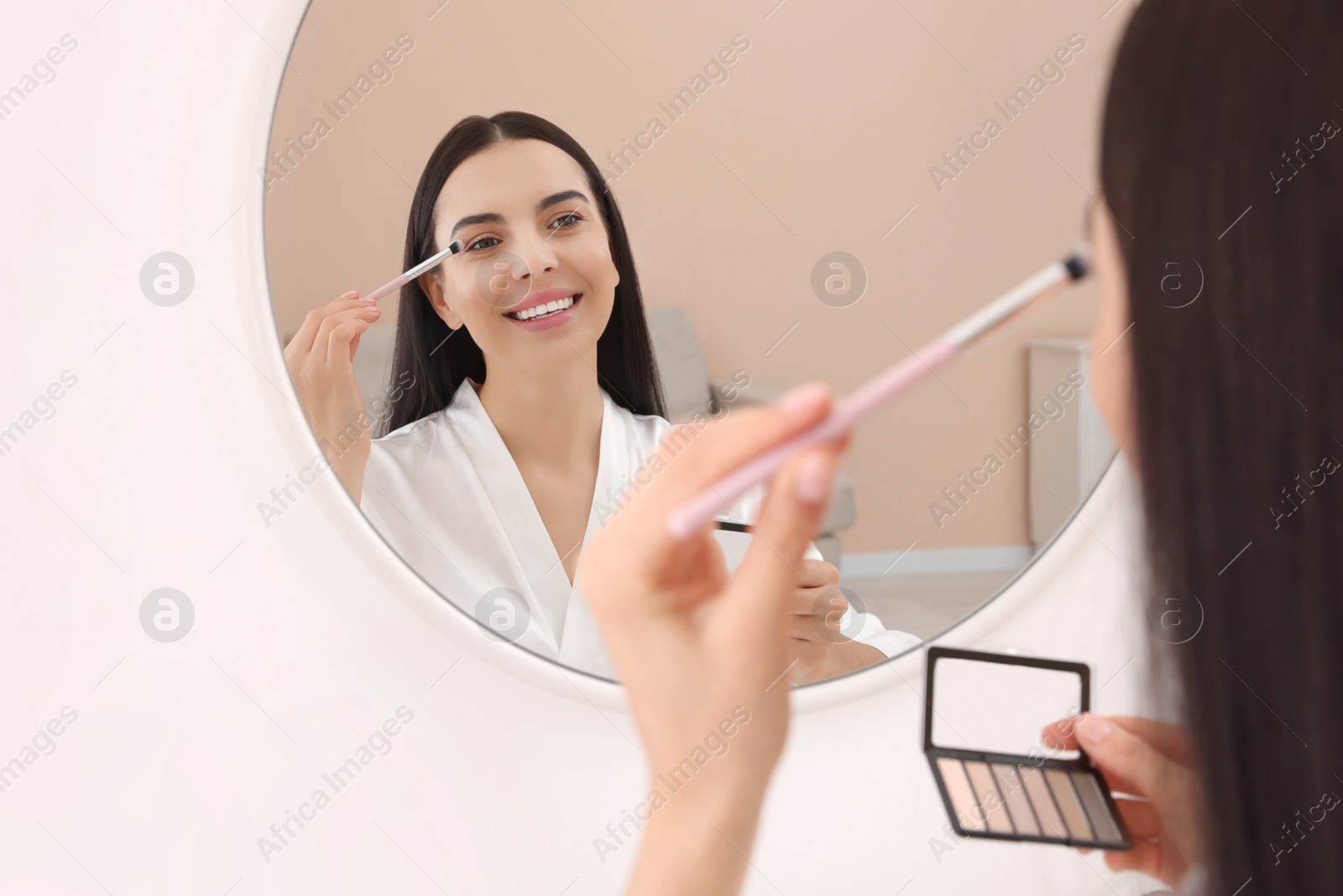 Photo of Beautiful young woman applying eyeshadow with brush near mirror indoors